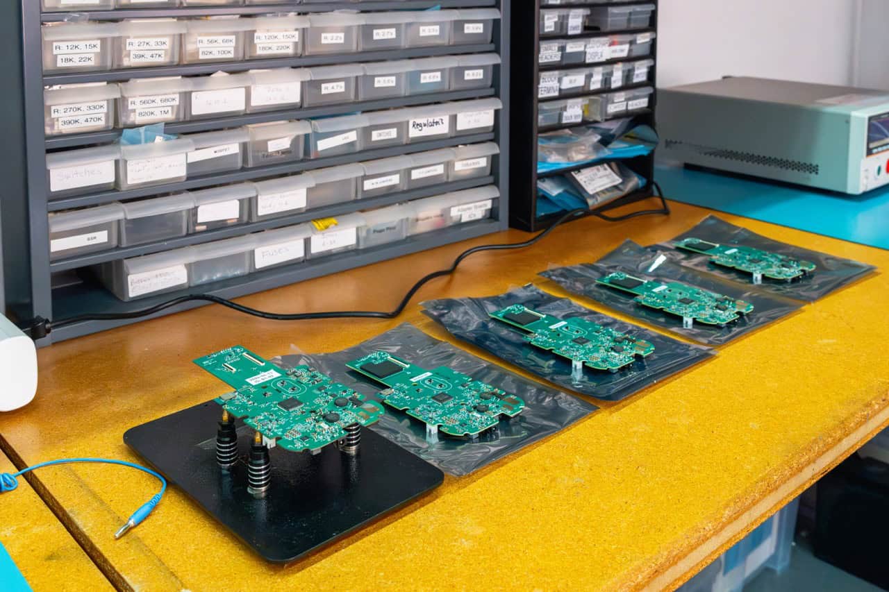 DFM test setup: PCBs on anti-static bags beside a spring-loaded test jig on a work bench; parts drawers in the background.