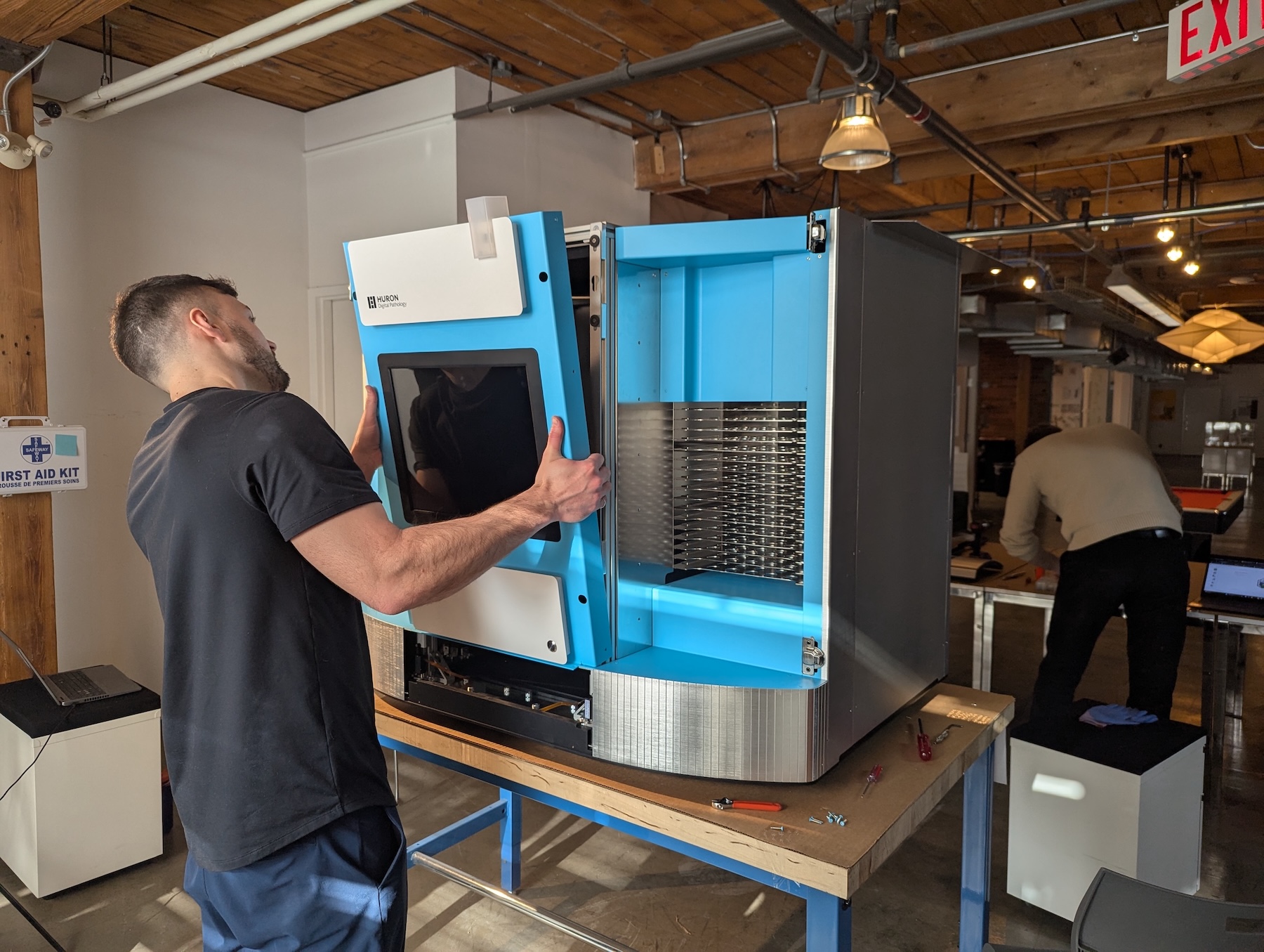 Cortex Design technician installing or removing the front panel of a Huron Digital Pathology medical device, showing internal components and slide storage racks in a workshop setting.