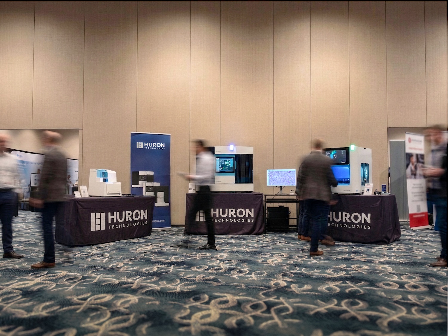 Huron Digital Pathology trade show booth displaying medical devices with branded tables and attendees walking by.
